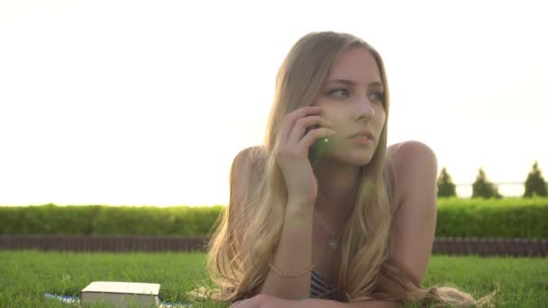 Young beautiful girl is lying on the lawn and talking on the phone - Footage, Video