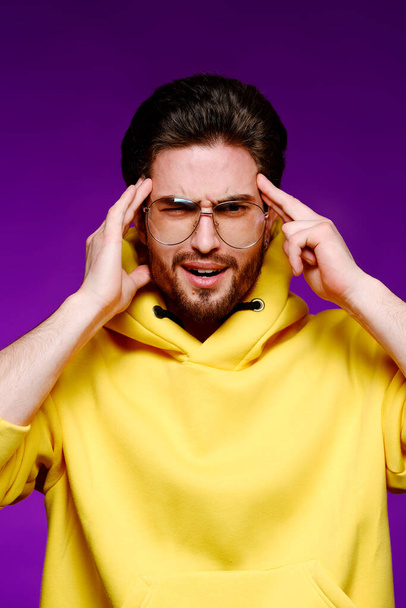 A young man of 25-30 years in glasses and a yellow sweatshirt emotionally poses holding his head on a purple background. - Photo, Image