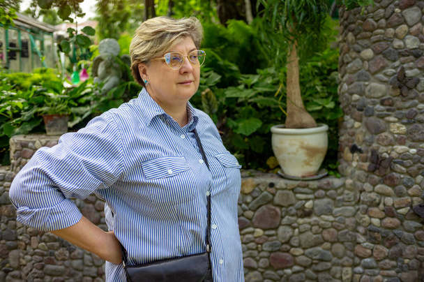 Μια ώριμη γυναίκα με ριγέ πουκάμισο και γυαλιά κοιτάζει από απόσταση. Ξεκουράζεται στο πάρκο της πόλης - Φωτογραφία, εικόνα