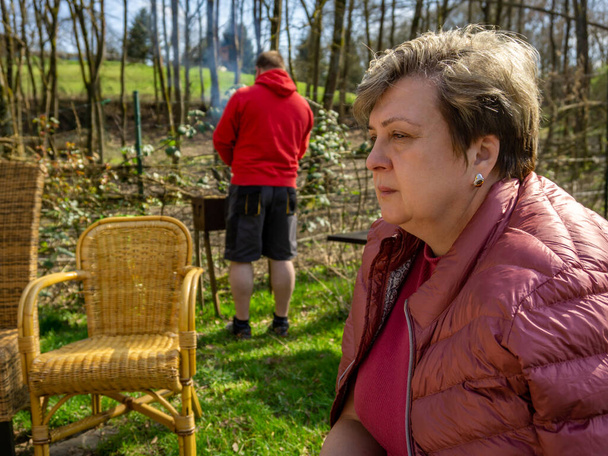 A Mature sad woman in a jacket is resting in a city Park and looking into the distance. In the background there is a guy in a red jacket doing a barbecue - Zdjęcie, obraz