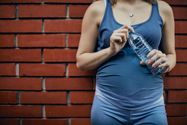 Close-up de esportista grávida tocando barriga, segurando garrafa de água e descansando após o exercício
. - Foto, Imagem