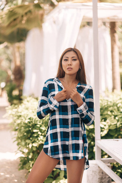 Street portrait of a young brunette asian woman in a plaid dress standing and posing in the city park outdoor, with blurred palm trees background. - 写真・画像