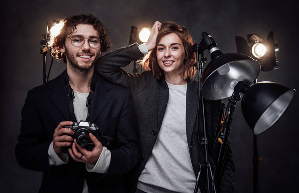 Команда из двух фотографов держит цифровую камеру и световое оборудование, позирующее в студии - Фото, изображение