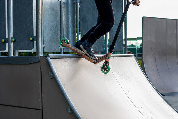 Tiener jongen rijdt op kick scooter in outdoor skatepark. Extreme zomersporten buiten. Voeten van mannelijke tiener 10 jaar oud rijden op. Leer trucjes. Sport als levensstijl - Foto, afbeelding