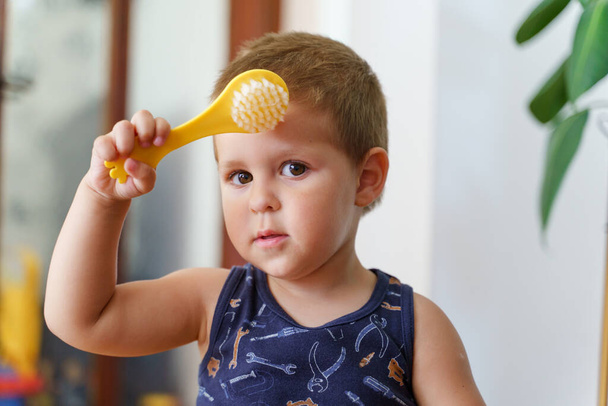 Porträt eines kleinen kaukasischen Jungen, der zu Hause eine Haarbürste hält - kleines Kind, zwei Jahre alt, steht im Zimmer und blickt tagsüber in die Kamera - Konzept des Erwachsenwerdens - Foto, Bild