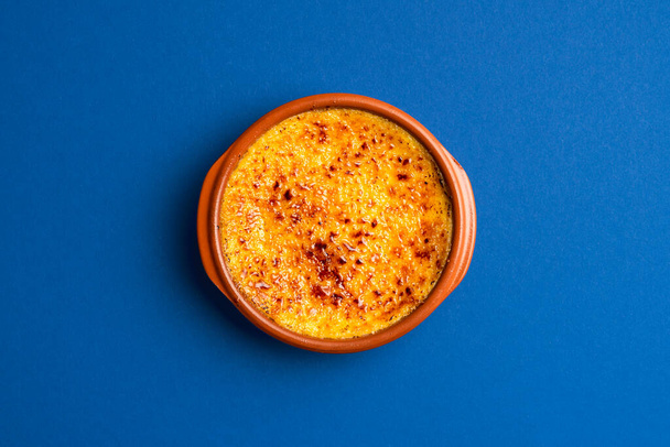 Drüber Ansicht mit frischer Creme Brulee auf blauem Hintergrund. Verbrannte Sahne mit karamellisiertem Zucker in einem Tonblech von oben. Französische Küche mit gebrannter Sahne. - Foto, Bild