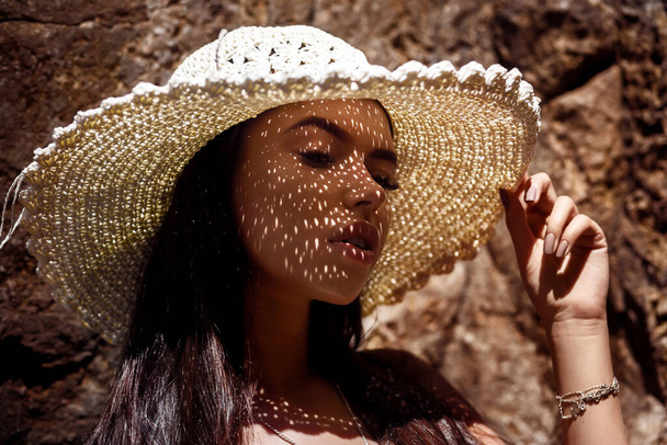 Closeup fashion portrait: a girl in a straw hat and with shadows on her face relaxes closing eyes - Fotó, kép