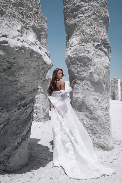 The tanned slim young woman with make up and dark hair wearing long white cape posing near white rocks. Back view, - Foto, Bild