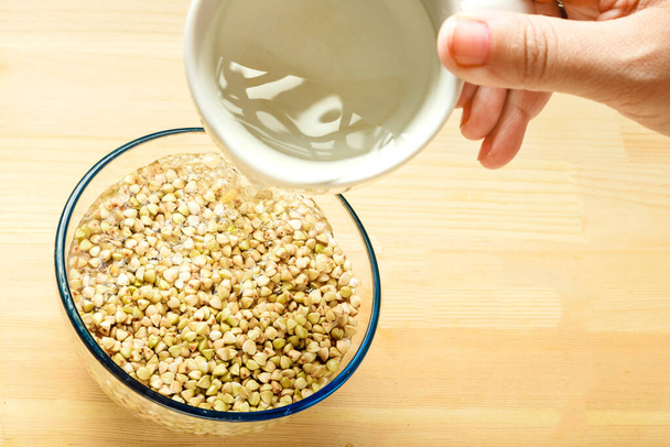 Female hand pours green buckwheat in a bowl with water from a cup. - Foto, afbeelding