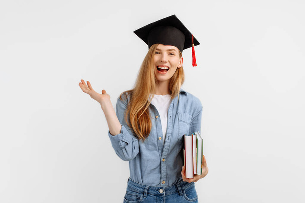 Happy excited happy beautiful girl graduate with a graduation hat on her head, holding books and having fun standing on a white background. Graduation, University graduation - Zdjęcie, obraz