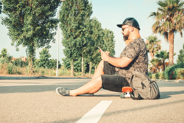 Uomo curvy con pantaloni corti che chiacchierano con il telefono seduto su uno skateboard. - Foto, immagini