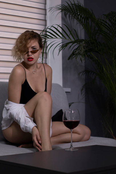 Sexy, beautiful blonde girl in a black bodysuit, a white shirt and sunglasses with a glass of red wine on a gray sofa - Фото, зображення