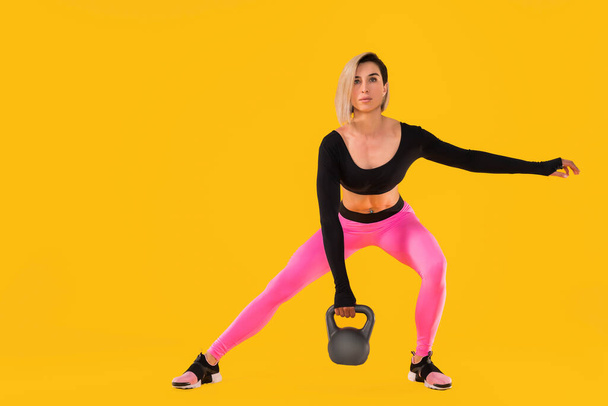Fitness kobieta w modnej różowej i czarnej odzieży sportowej ćwiczyć z kettlebell na żółtym tle. - Zdjęcie, obraz