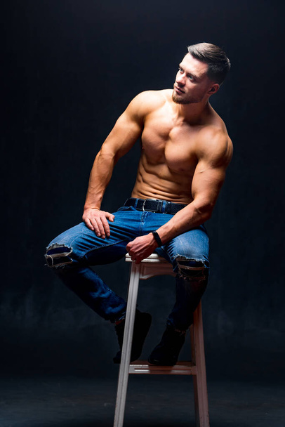 Man with perfect body sitting on high chair. Dark background. Male beauty. - Фото, изображение