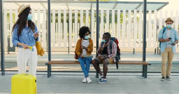 Smíšené rasy mladých mužů a žen v lékařských maskách stojící na autobusové zastávce a čekající na transport. Multietničtí turisté na vlakovém nádraží. Koncept cestování. - Záběry, video