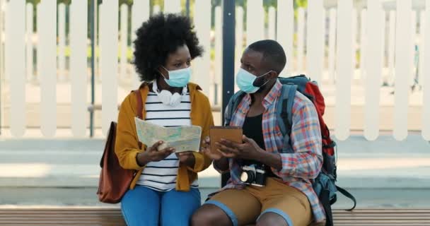 Αφροαμερικανοί νέοι τουρίστες με ιατρικές μάσκες κάθονται στον πάγκο στη στάση του λεωφορείου και περιμένουν τη μεταφορά τους. Όμορφη γυναίκα με χάρτη της πόλης και όμορφος άντρας με συσκευή tablet ψάχνει για διαδρομή. - Πλάνα, βίντεο