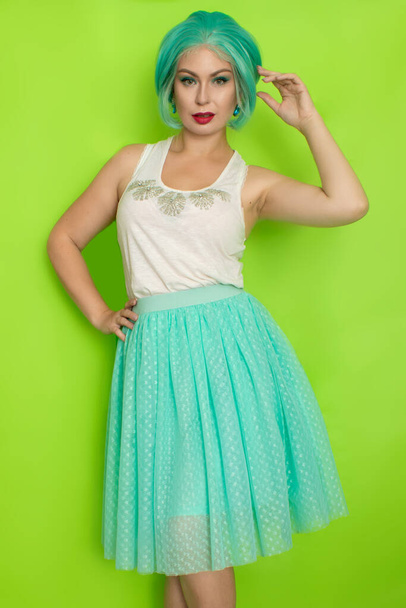 Πορτρέτο μιας όμορφης νεαρής γυναίκας με κοντά μπλε μαλλιά σε ένα στούντιο σε πράσινο φόντο. Μεσαίου μήκους ρομαντική φούστα και λευκή μπλούζα - Φωτογραφία, εικόνα