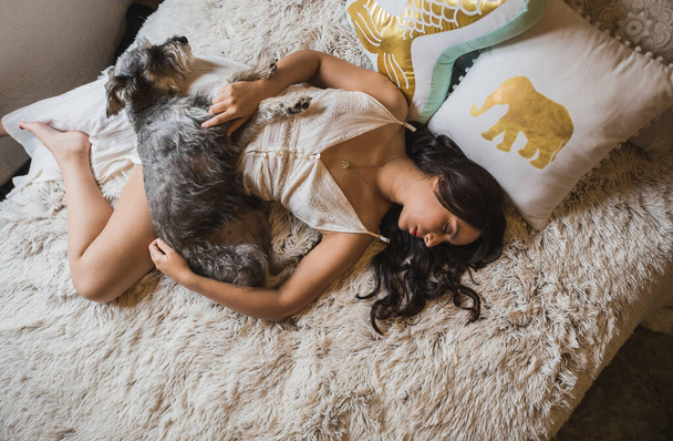 Girl lying down, resting and taking a nap, dressed in a white blanket dress, in her bed next to her Schnauzer dog, an afternoon of relaxation and company of her pet. - Photo, Image