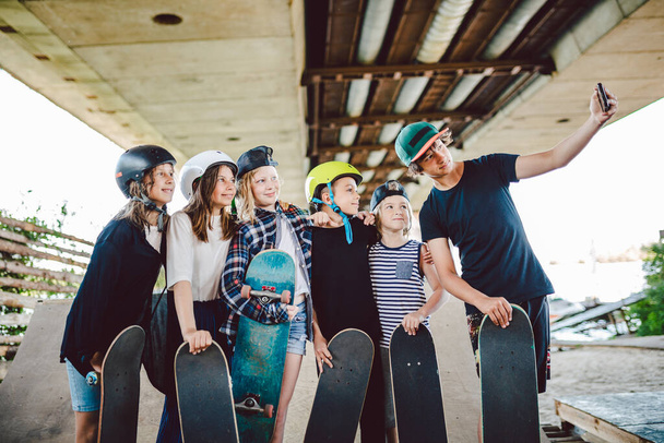 grupa skateboardzistów dzieci tworzą treści w mediach społecznościowych przez telefon podczas spędzania czasu na rampie w skateparku. Przyjaciele stoją razem z deskorolkami przytulając i robiąc zdjęcie selfie na smartfonie. - Zdjęcie, obraz