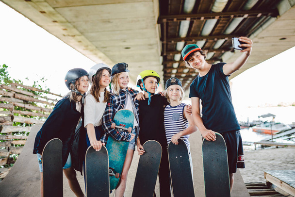 Gruppe von Skateboardern Kinder machen Social-Media-Inhalte am Telefon, während sie Zeit auf der Rampe im Skatepark verbringen. Freunde stehen mit Skateboards zusammen und umarmen sich und machen Selfie-Fotos mit dem Smartphone. - Foto, Bild