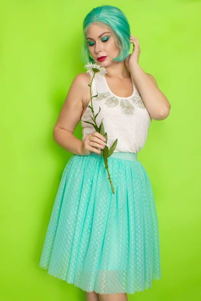 Studio portrait of beautiful mint hair feminine in white top and light blue skirt holding big chamomile in her hands, over green background - Photo, image
