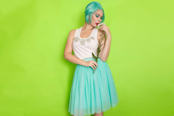 Studio portrait of beautiful mint hair feminine in white top and light blue skirt holding big chamomile in her hands, over green background - Photo, Image