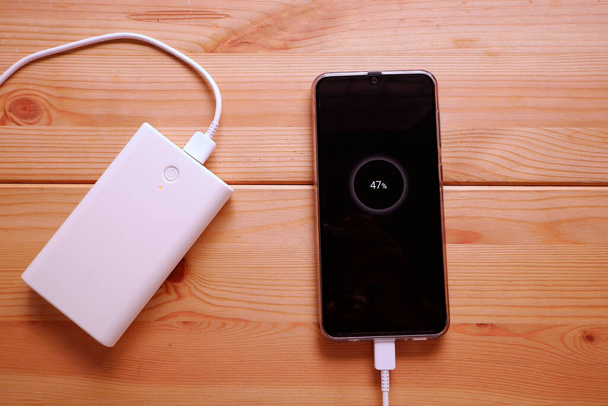 Smartphone se está cargando con batería de reserva o banco de energía blanca en la mesa de madera de escritorio
 - Foto, Imagen