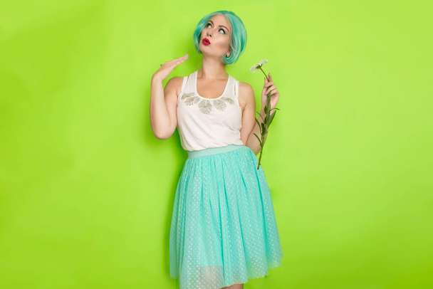 Studio portrait of beautiful mint hair feminine in white top and light blue skirt holding big chamomile in her hands, over green background - Foto, immagini