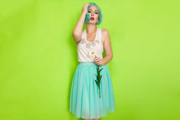 Studio portrait of beautiful mint hair feminine in white top and light blue skirt holding big chamomile in her hands, over green background - Foto, Imagen