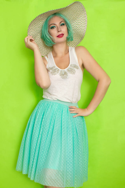 Beautiful young mint haired woman in white top and blue middle skirt wearing white and black hat. Studio photo over green background - Φωτογραφία, εικόνα