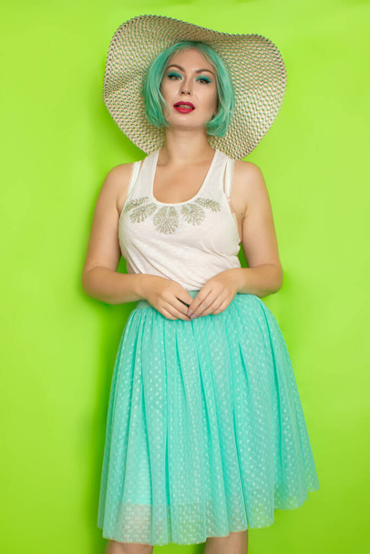 Beautiful young mint haired woman in white top and blue middle skirt wearing white and black hat. Studio photo over green background - Zdjęcie, obraz