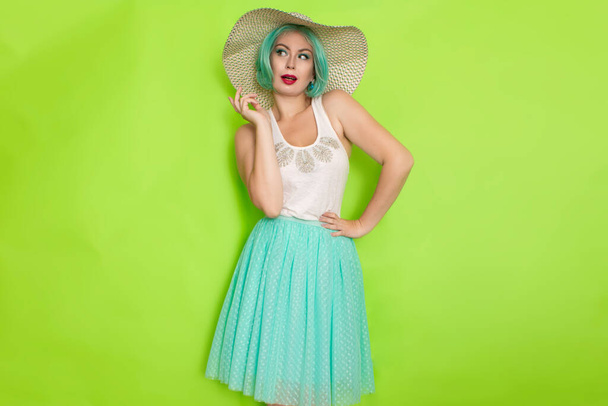 Beautiful young mint haired woman in white top and blue middle skirt wearing white and black hat. Studio photo over green background - Foto, imagen