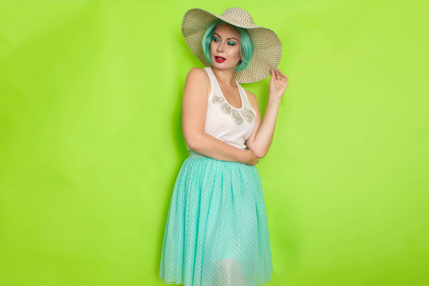 Beautiful young mint haired woman in white top and blue middle skirt wearing white and black hat. Studio photo over green background - Foto, imagen