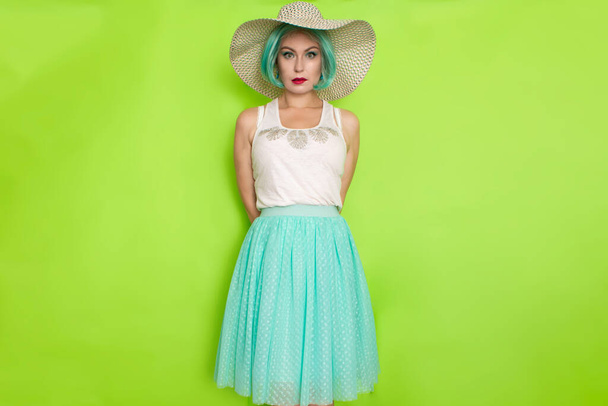 Beautiful young mint haired woman in white top and blue middle skirt wearing white and black hat. Studio photo over green background - Фото, изображение