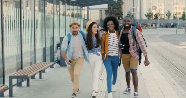 Jonge vrolijke Kaukasische en Afro-Amerikaanse toeristen die plezier hebben in de buitenlucht, rennen en springen. Vreugdevolle gemengde rassen mannen en vrouwen die samen reizen. In de stad bij de bushalte. Meertaligheid. - Video