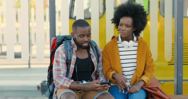 Αφροαμερικανοί νέοι τουρίστες που κάθονται στον πάγκο στη στάση του λεωφορείου, μιλώντας και περιμένοντας για μεταφορά. Όμορφη γυναίκα και όμορφος άντρας ψάχνουν τρόπο για το smartphone και το σχεδιασμό της διαδρομής. Ταξιδιώτες. - Πλάνα, βίντεο