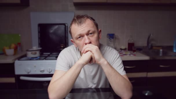 Een depressieve man zit 's avonds aan de keukentafel, kijkt direct naar de camera, camerabeweging - Video