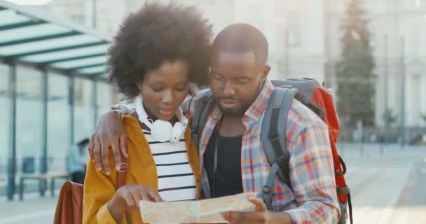 Portret van gelukkige jonge Afro-Amerikaanse paar toeristen staan op straat bij de bushalte en kijken naar de kaart. Mooie vrouw en knappe man plannen route met stadsplan. Reisconcept. - Video