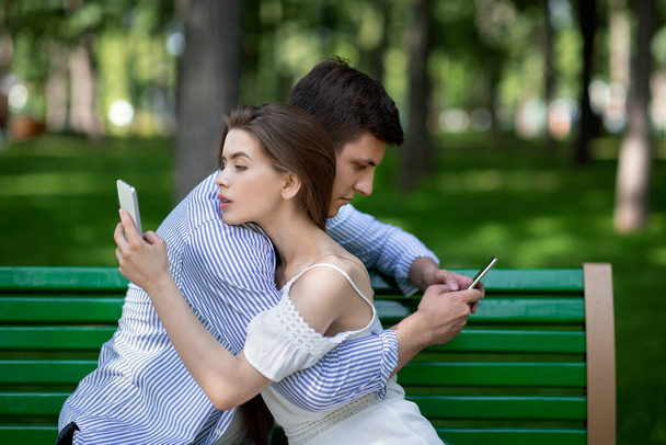 Gadget coppia dipendente utilizzando i telefoni cellulari mentre abbraccia sulla panchina al parco - Foto, immagini