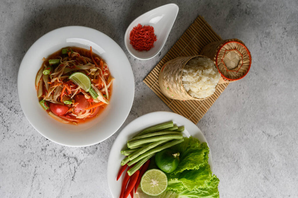 Ταϊλανδέζικη σαλάτα παπάγια σε λευκό πιάτο με κολλώδες ρύζι σε ψάθινο καλάθι μπαμπού και αποξηραμένες γαρίδες. - Φωτογραφία, εικόνα