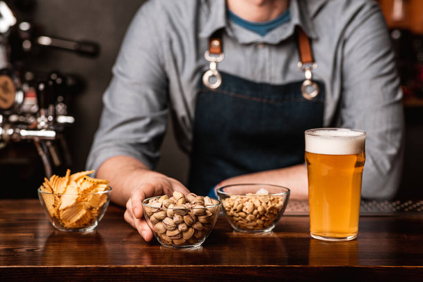 Хрустящие закуски от бармена. Фисташки, орехи и чипсы в тарелках и стакан светлого пива с пеной
 - Фото, изображение
