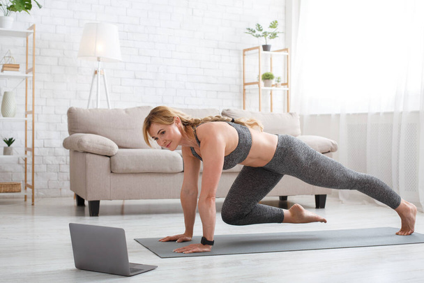 Fitness workout of sporty woman. Exercises for legs on mat with laptop in living room interior - Photo, Image