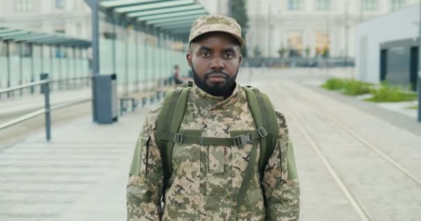 Retrato de um jovem soldado afro-americano bonito de boné com mochila na rua. Homem militarista olhando para a câmera na estação de trem. Paragem de autocarro. Uniforme militar. Aumento de zoom. - Filmagem, Vídeo