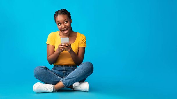 Позитивна африканська дівчинка використовує смартфон сидячи над синім фоном, панорама - Фото, зображення