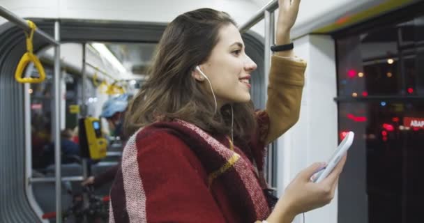 Blanke jonge mooie vrouw in jas, sjaal en koptelefoon luisteren naar muziek op de smartphone en 's avonds thuiskomen. Aantrekkelijke stijlvolle vrouwelijke passagier in tram en telefoon. - Video