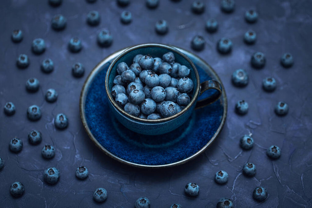 Bleuets frais dans une tasse en céramique bleue avec une soucoupe sur un fond sombre, une belle tasse de café remplie de baies sauvages - Photo, image
