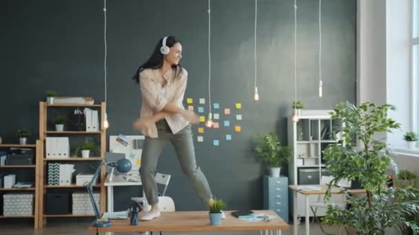 Happy young woman dancing on office table wearing headphones having fun alone - Filmmaterial, Video