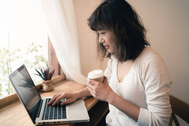 Young Asian woman working online with laptop computer and drinking a cup of coffee. A woman with smiley face working from home. Social distancing concept. Bangkok, Thailand - Φωτογραφία, εικόνα