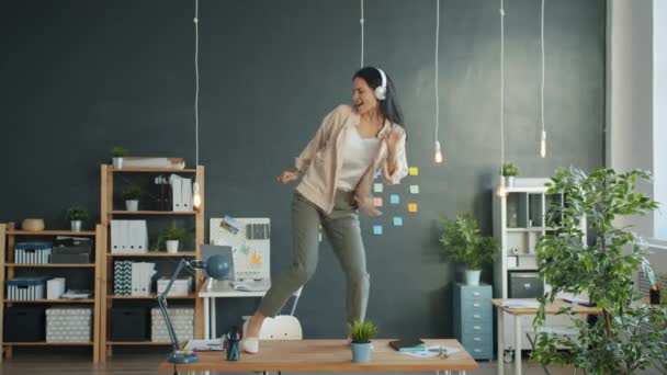 Beautiful girl office worker dancing on desk in office wearing headphones enjoying break - Filmagem, Vídeo
