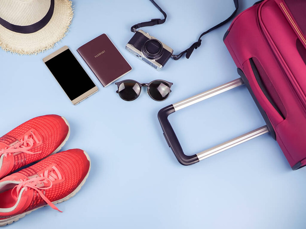 Tasainen lay matkustajan tarvikkeet, passi, hattu, matkapuhelin, kamera, aurinkolasit, lenkkarit ja vaaleanpunainen matkalaukku sinisellä pohjalla. matkustuskäsite. - Valokuva, kuva